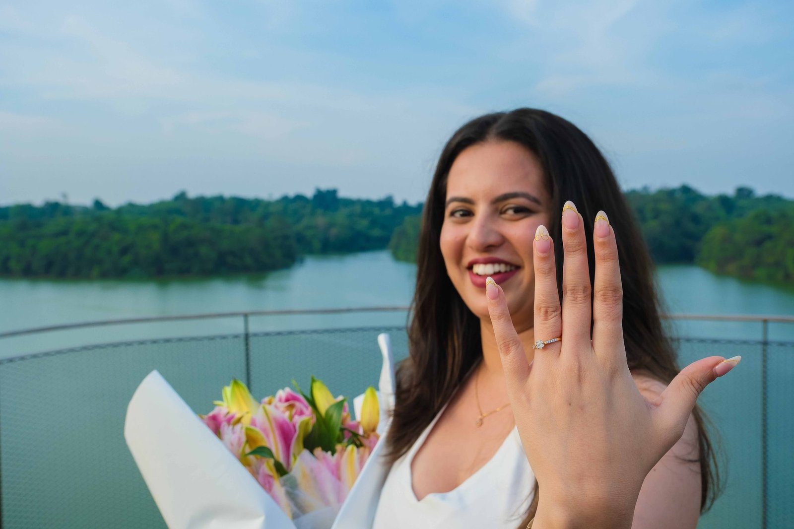 A woman laughing joyfully while showing off her engagement ring beside a lake